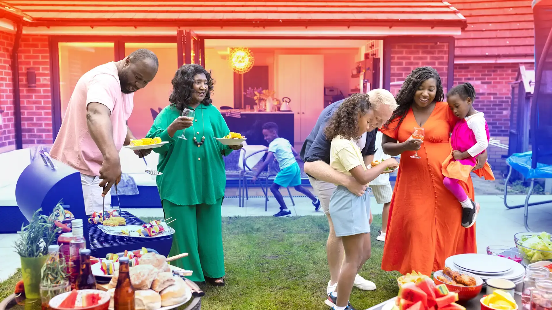 Multigenerational family enjoying a backyard barbecue on a sunny day. People are serving food, holding drinks, and smiling while children play nearby.