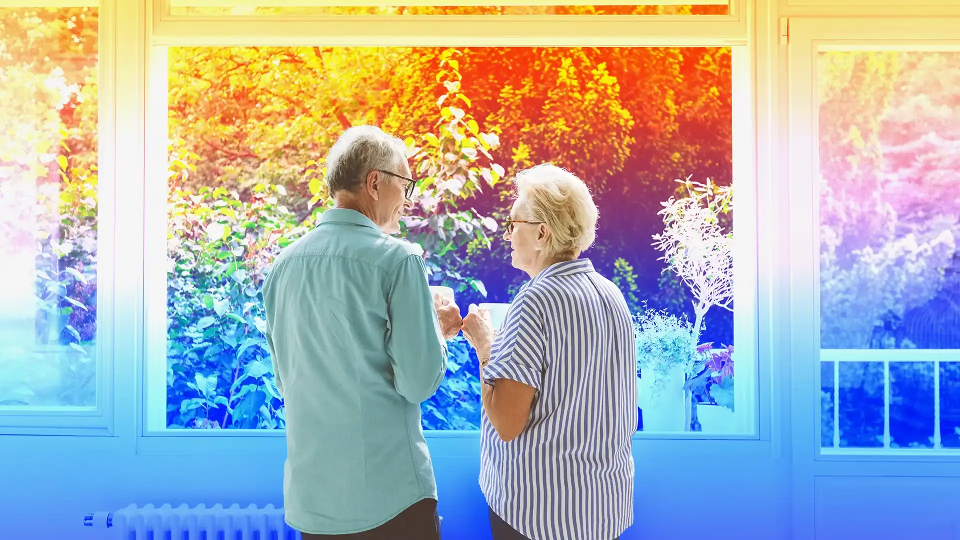 Older couple holding coffee mugs and smiling at each other while standing by a large window.