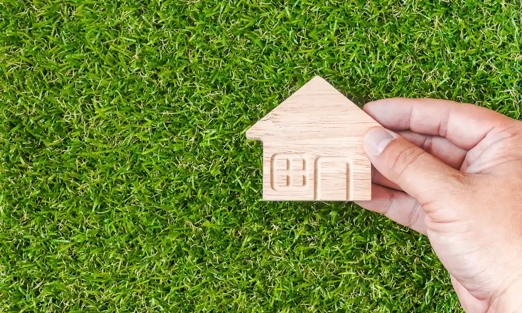 A hand holding a little 2D wooden house over astro turf