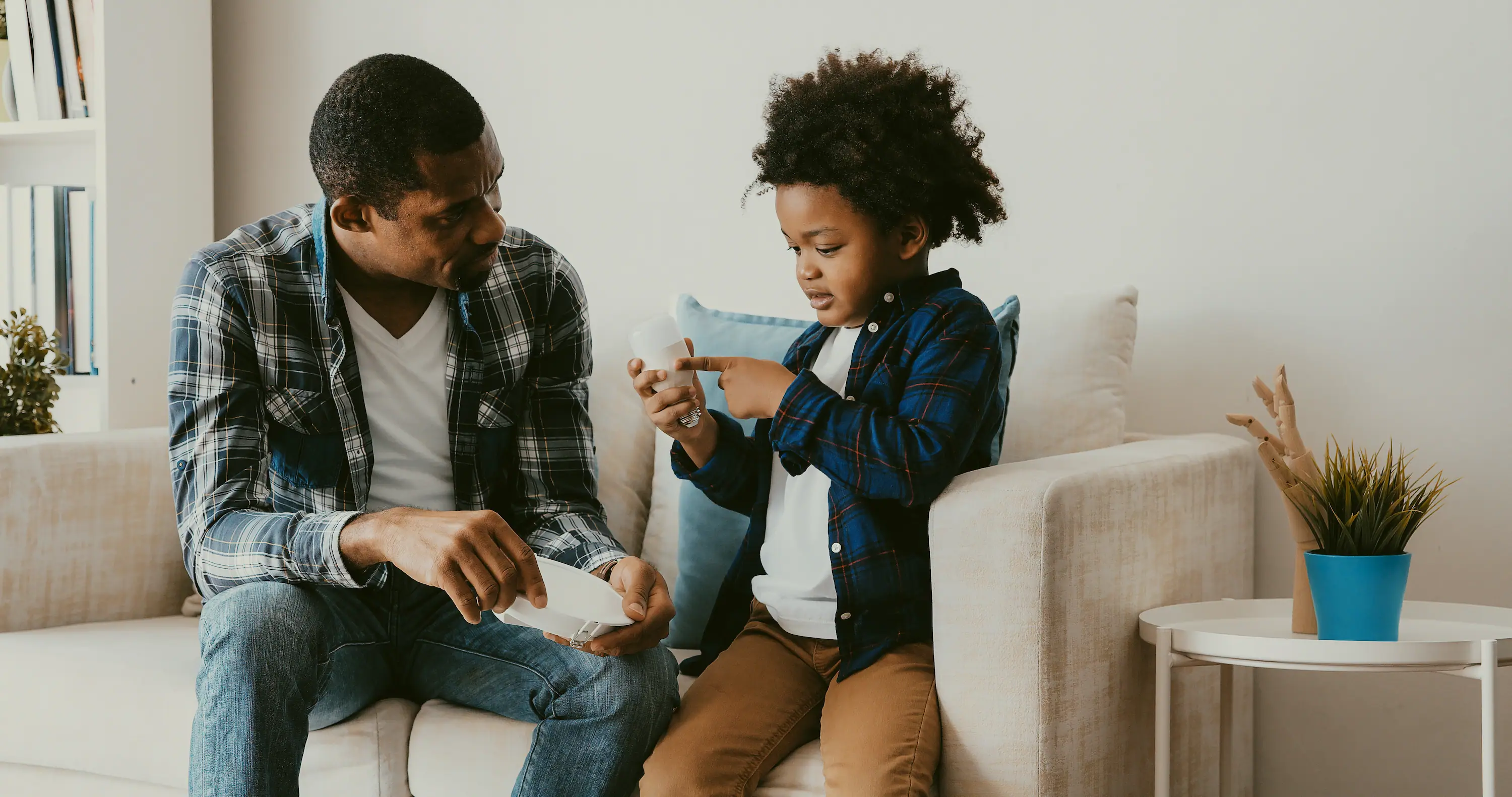 Father and young son sitting on a couch, learning how to use a smart home device together.