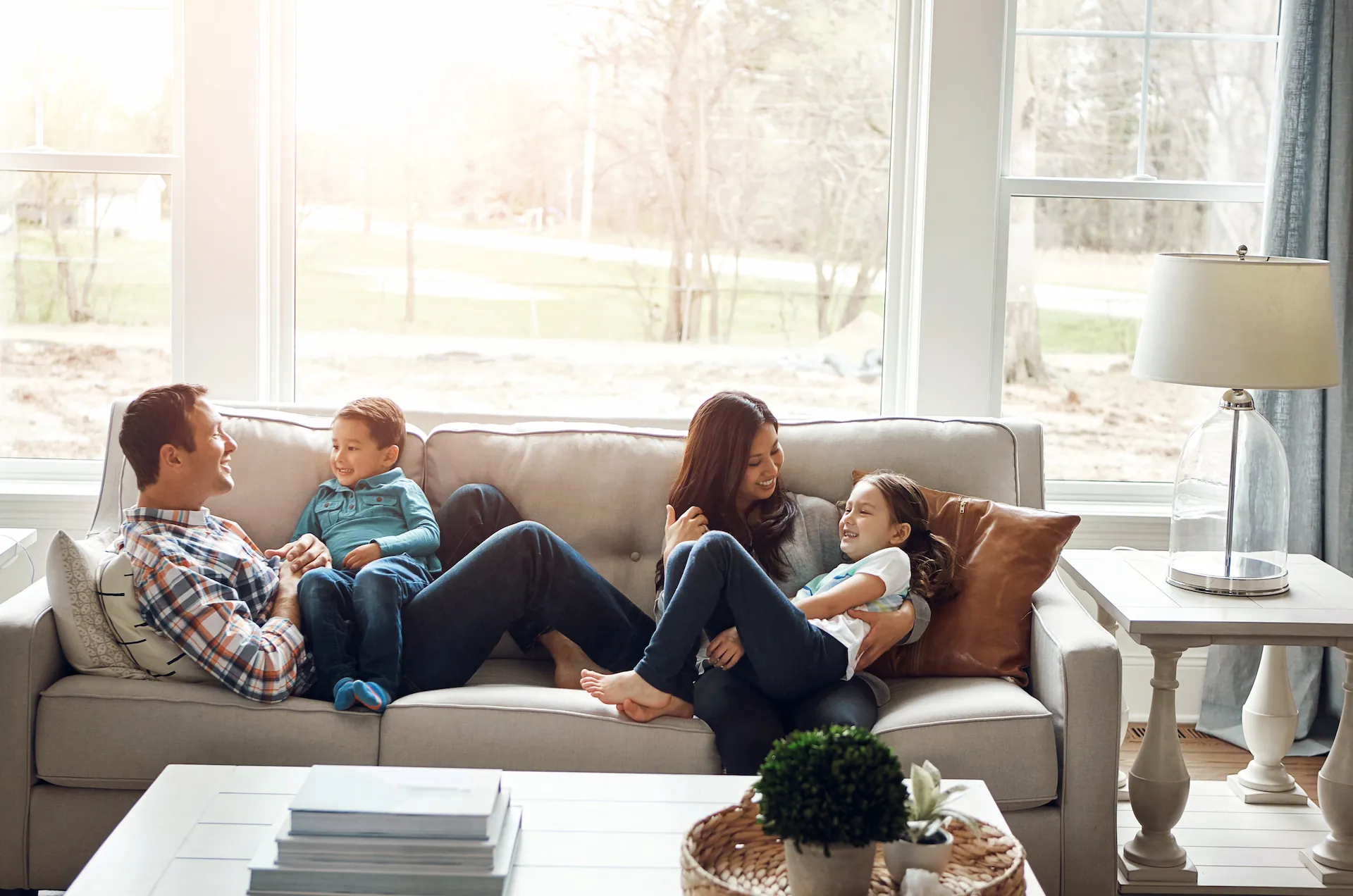 Family of four relaxing together on a couch in a bright living room, smiling and laughing near large windows that let in natural light.