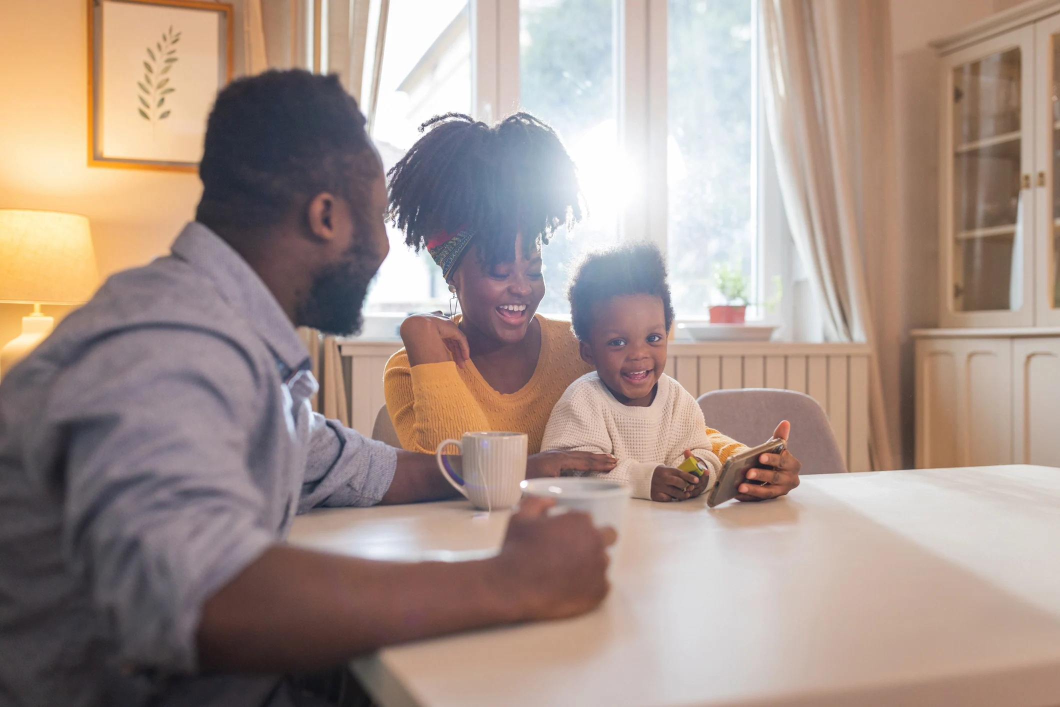 Family sitting at a dining table in a bright kitchen, smiling as a young child holds a smartphone while parents look on.