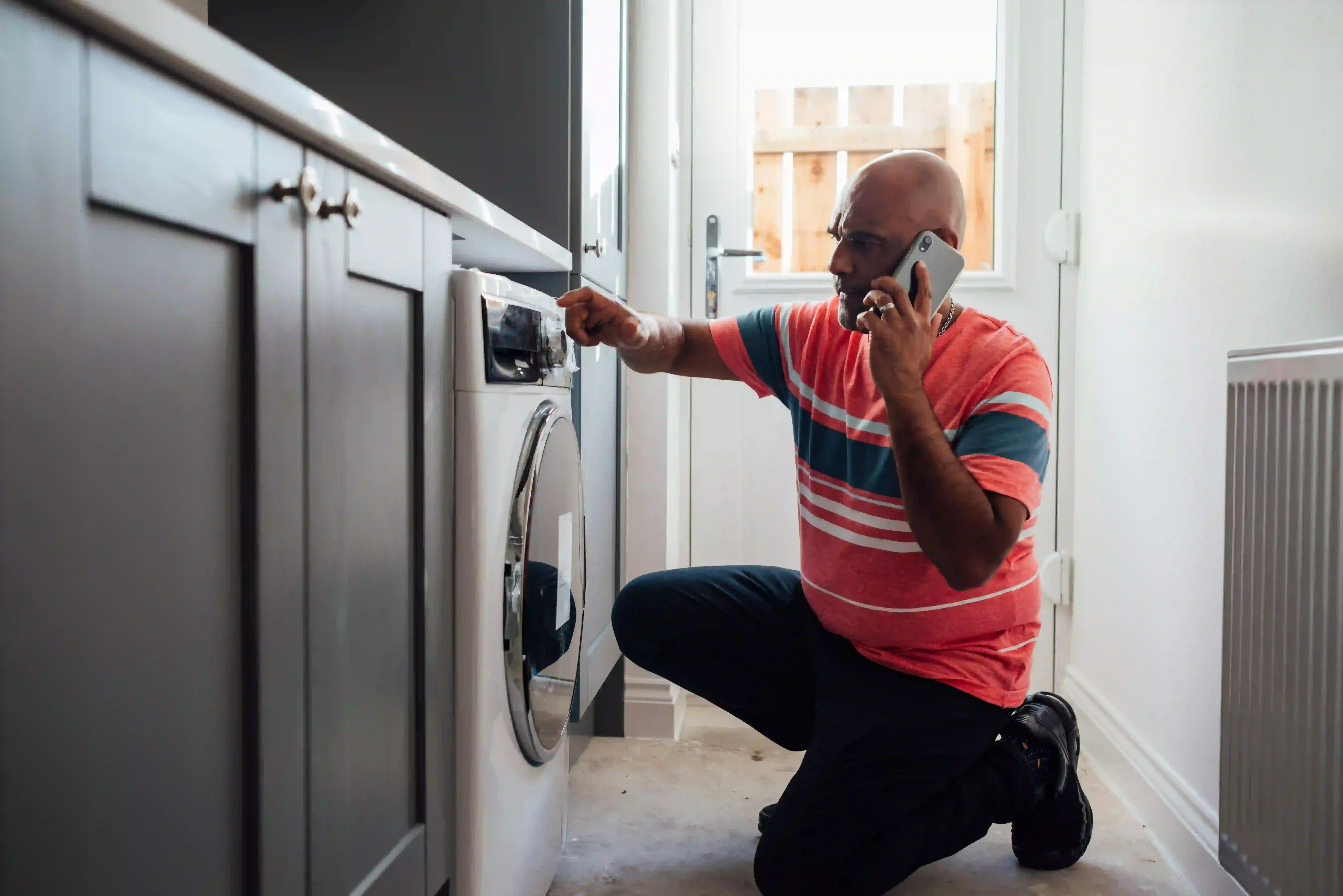 Man kneeling in front of a washing machine while talking on the phone, possibly troubleshooting an issue.