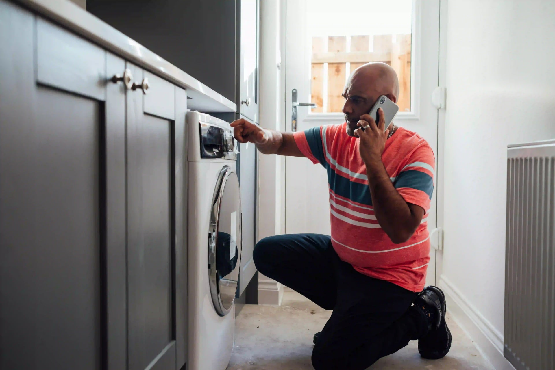 Man kneeling in front of a washing machine while talking on the phone, possibly troubleshooting an issue.