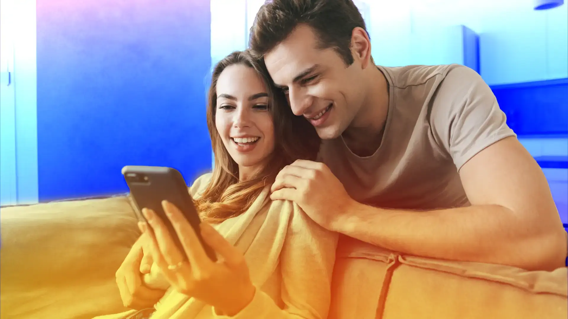 Smiling couple sitting on a couch at home, looking at a smartphone together.