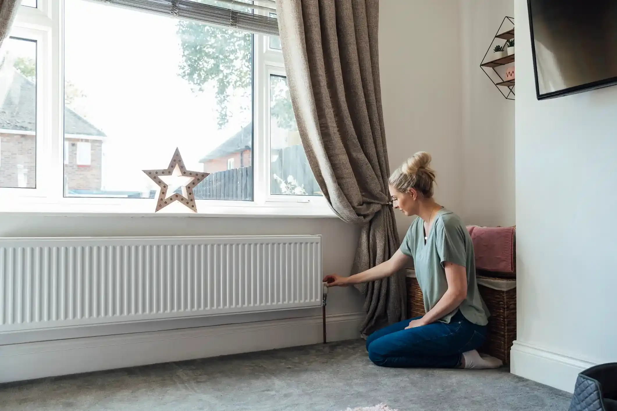 Woman kneeling in a living room adjusting the valve on a home radiator beneath a window.